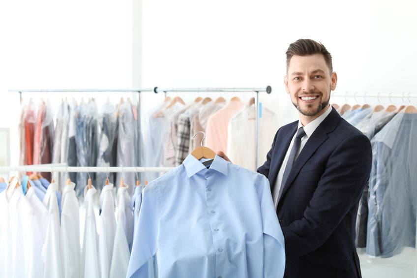 A man in a suit holding up a blue shirt in a room filled with various shirts on a rack.