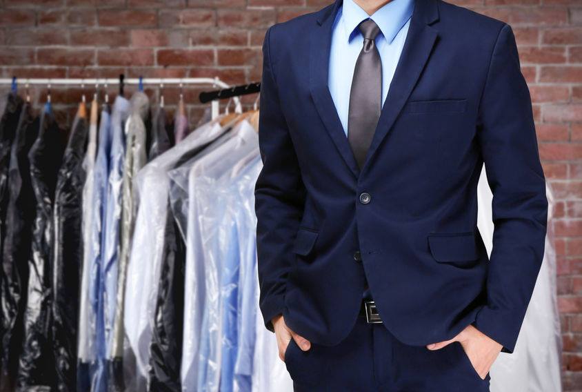 A well-dressed man in a blue suit and tie stands in front of a rack of clothing, showcasing a professional and polished appearance.